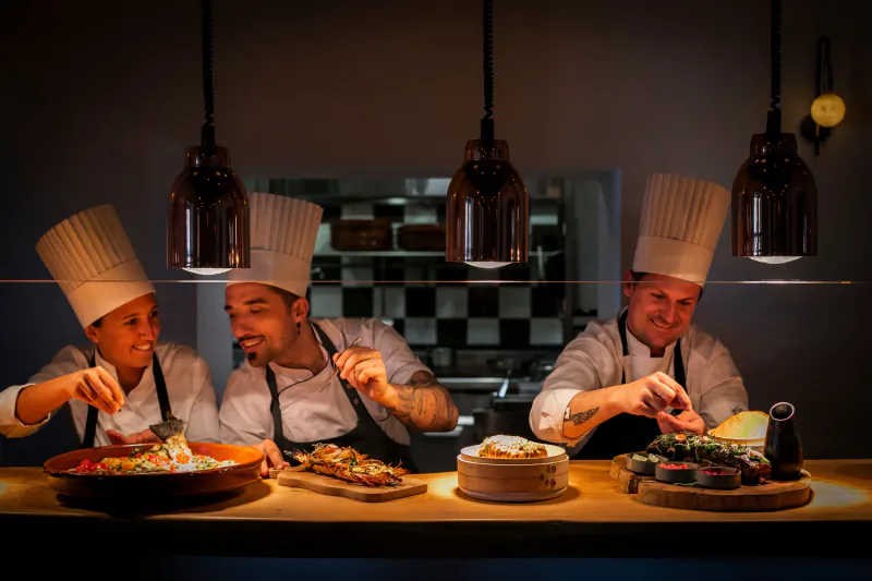 Three smiling chefs in white uniforms and hats plating colorful dishes behind a restaurant counter under pendant lights
