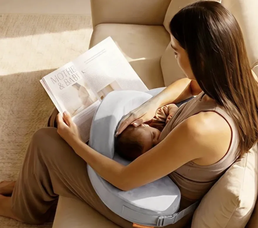 Mother sitting in armchair, reading magazine while baby sleeps on blue nursing pillow in her lap