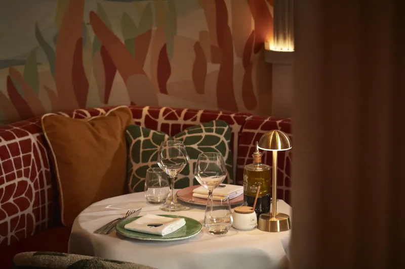 Elegant restaurant table set with white linens, green plates, wine glasses, and a brass lamp against a colorful abstract mural