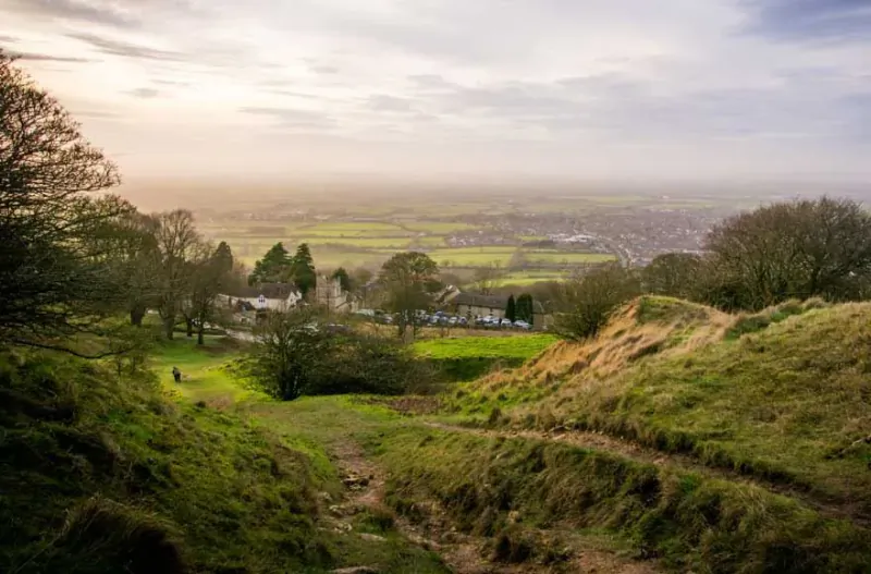 Panoramic view of Moreton-in-Marsh valley with rolling green fields, scattered houses, and a winding hillside path at sunset