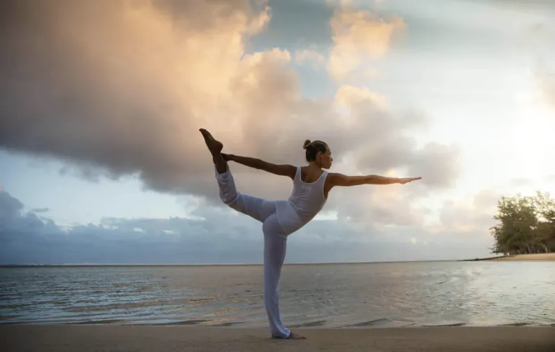 Woman in white yoga attire balances in dancer pose on a sandy beach at sunset with ocean and clouds behind