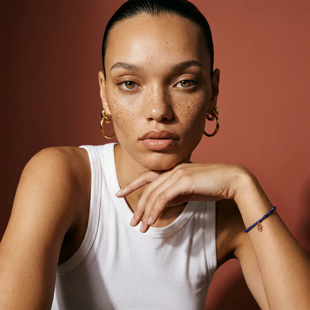 Close-up portrait of a woman with freckles wearing gold hoop earrings, a white tank top, and a blue beaded bracelet with a small charm