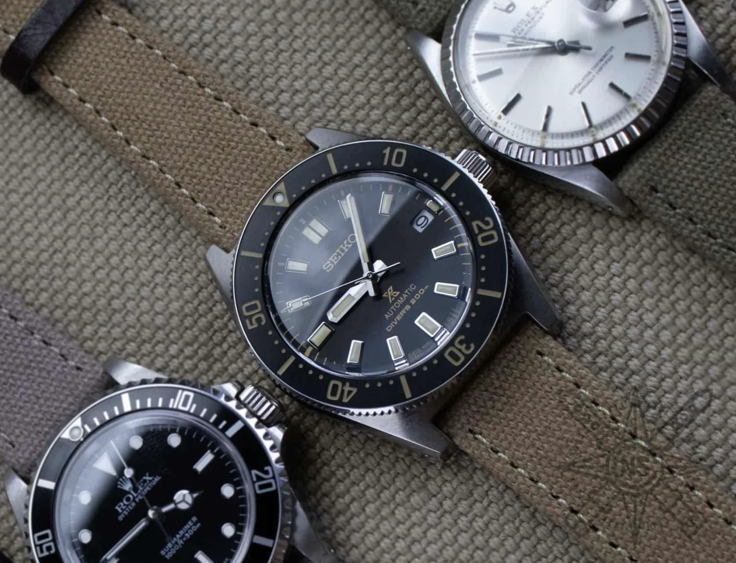 Three dive watches on olive canvas: Seiko 5 Sports black dial center, Rolex Submariner bottom left, Seiko white dial top right