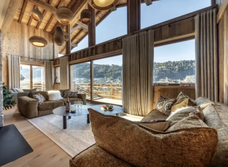 Bright modern living room with large windows, wooden beams, brown sofas, and mountain view