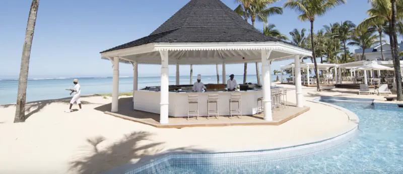 Beachfront bar with white thatched roof, staff behind counter, palm trees and ocean view at Heritage Le Telfair Mauritius