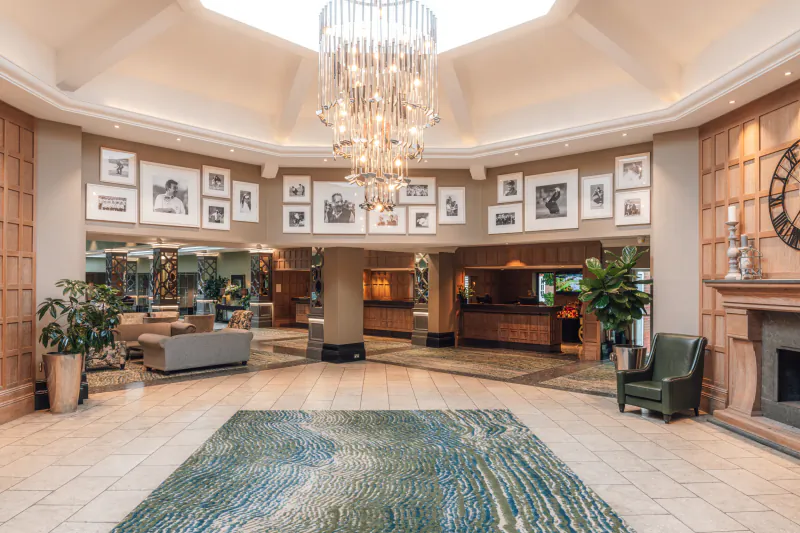 Luxurious hotel lobby with crystal chandelier, reception desk, framed photos on walls, green rug, and fireplace