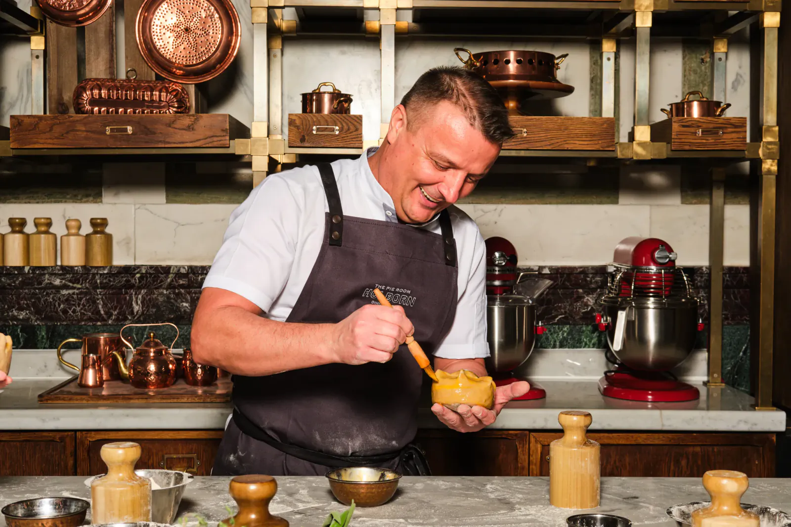 Chef in apron smiling while scooping yellow filling from a bowl with a spatula in a professional kitchen