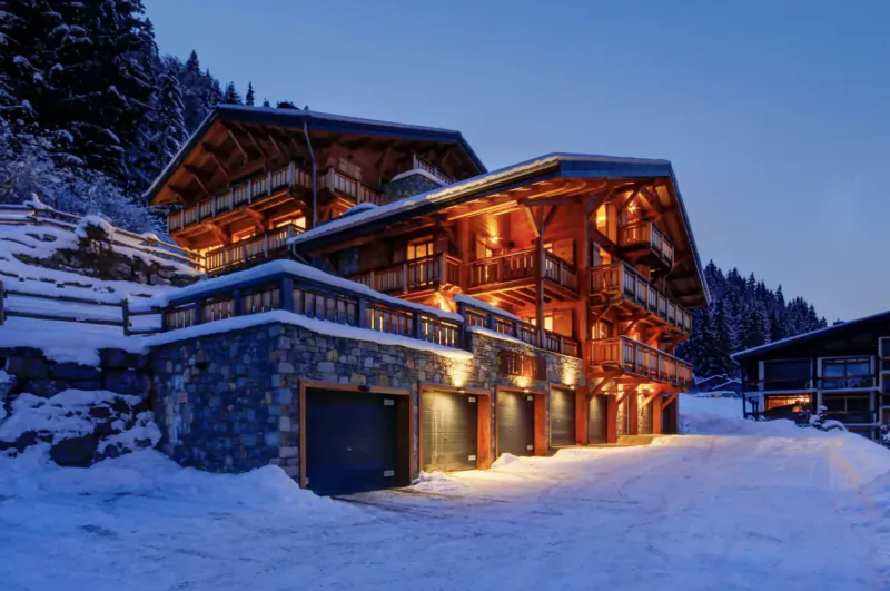 Snow-covered luxury wooden chalet with stone base, lit balconies, and garage doors at dusk