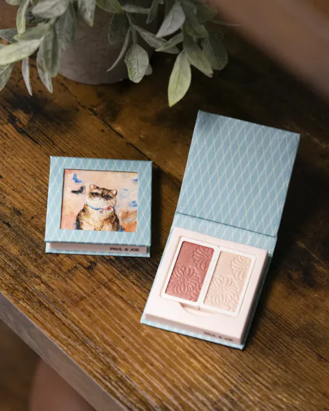 Open Paul & Joe makeup palette with cat illustration on lid, showing pink and white embossed powder pans on wooden table