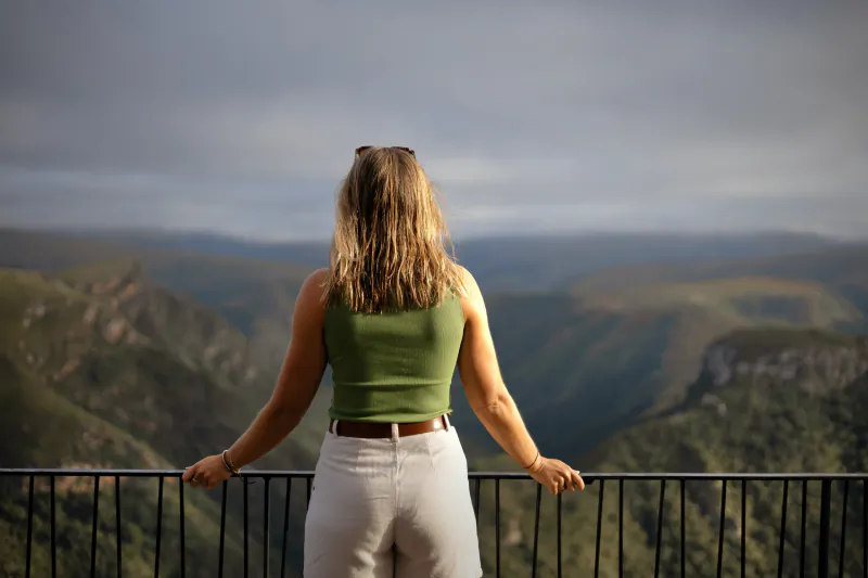 Woman in green top and white pants stands at railing overlooking misty mountain range
