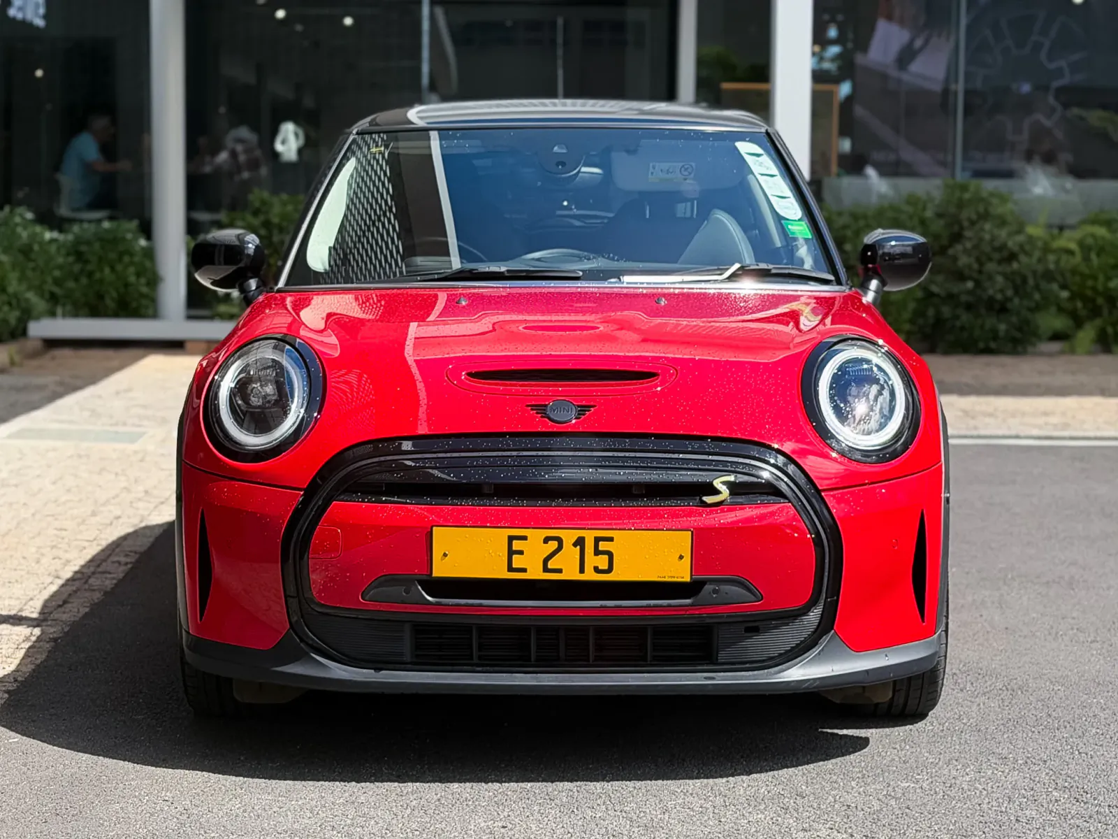 Front view of a glossy red Mini Cooper S with yellow license plate E 215 parked outside a dealership
