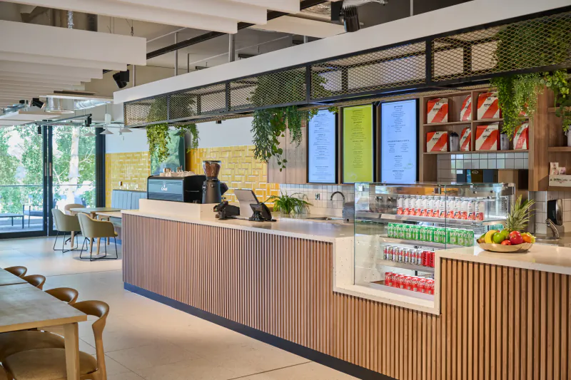 Modern hotel cafe counter with barista, wooden slat front, hanging plants, menu boards, and drinks fridge