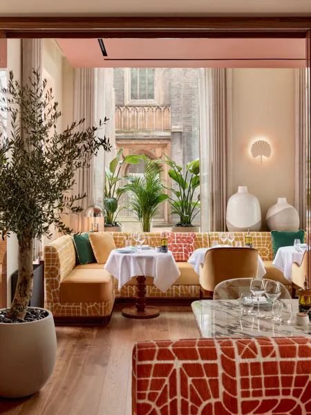 Elegant restaurant interior with mustard yellow patterned banquette seating, white tablecloths, and large potted olive tree near tall windows overlooking a building facade