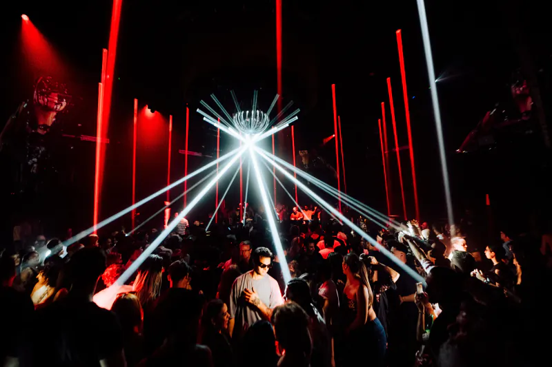 Crowd dancing under bright white and red laser beams at a dark nightclub with a central light fixture