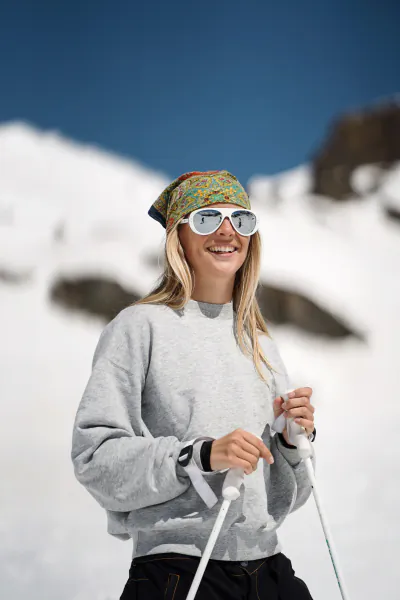 Smiling woman in grey sweatshirt and colorful headband holding ski poles on snowy mountain slope