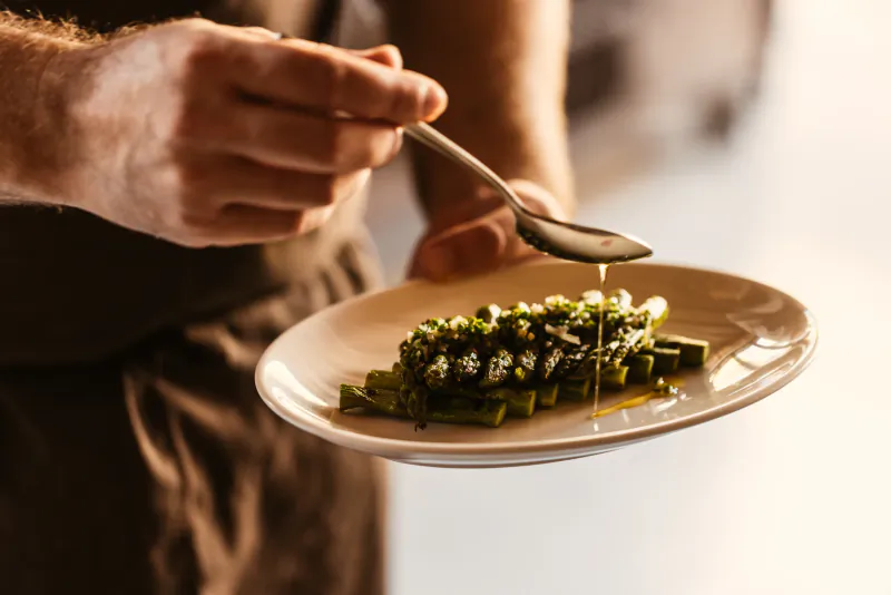 Chef drizzling sauce over plated green asparagus with herbs on a white plate
