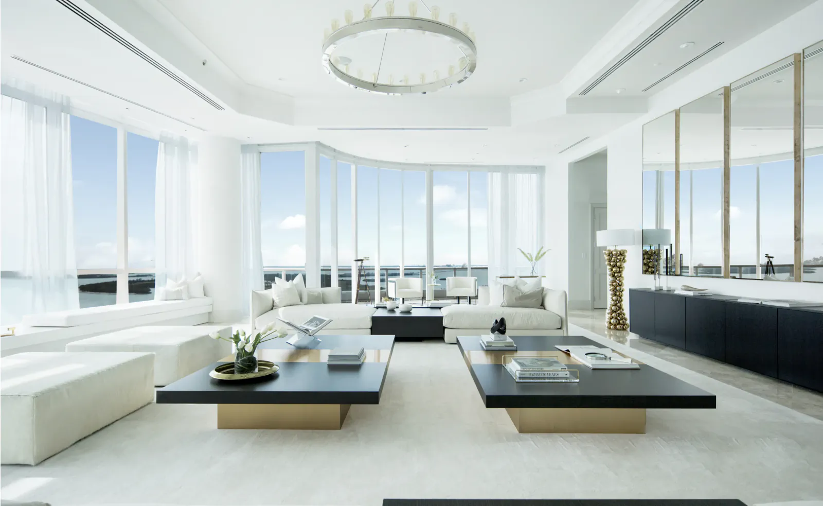 Bright modern living room with white sofas, black coffee tables, floor-to-ceiling windows overlooking the ocean, and a circular chandelier