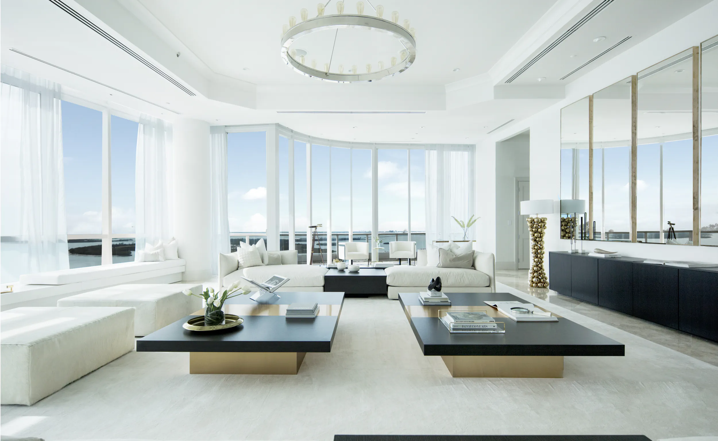 Bright modern living room with white sofas, black coffee tables, floor-to-ceiling windows overlooking the ocean, and a circular chandelier