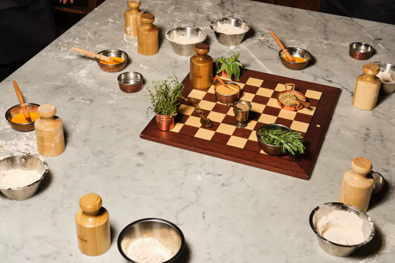 Overhead view of a marble table set for a pie masterclass with a wooden chessboard holding spices, herbs, and ingredients, surrounded by bowls of flour and wooden grinders