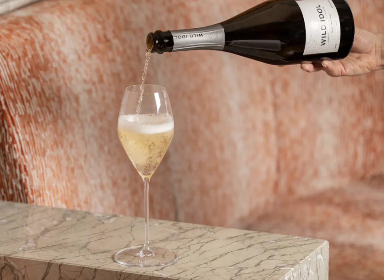 Hand pouring Wild Idol sparkling wine from a dark bottle into a flute glass on marble table