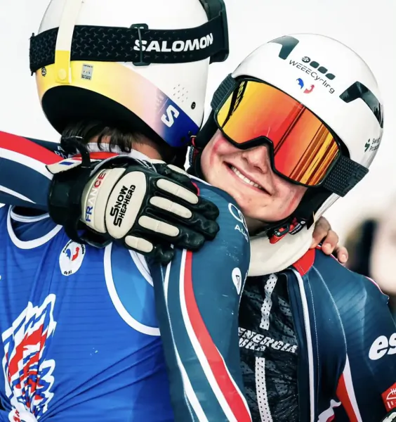 Two French snowboarders in Salomon helmets embrace, one smiling with orange goggles, both in blue-red suits
