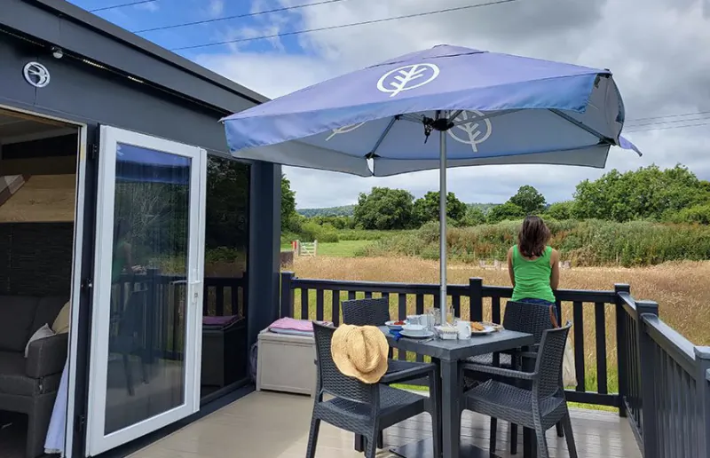 Woman in green top sits at outdoor table under blue umbrella on deck overlooking countryside at Moreton-in-Marsh camping site