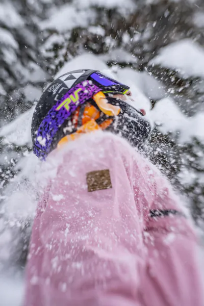 Snowboarder in pink jacket and black helmet with purple graphics, viewed from above in falling snow