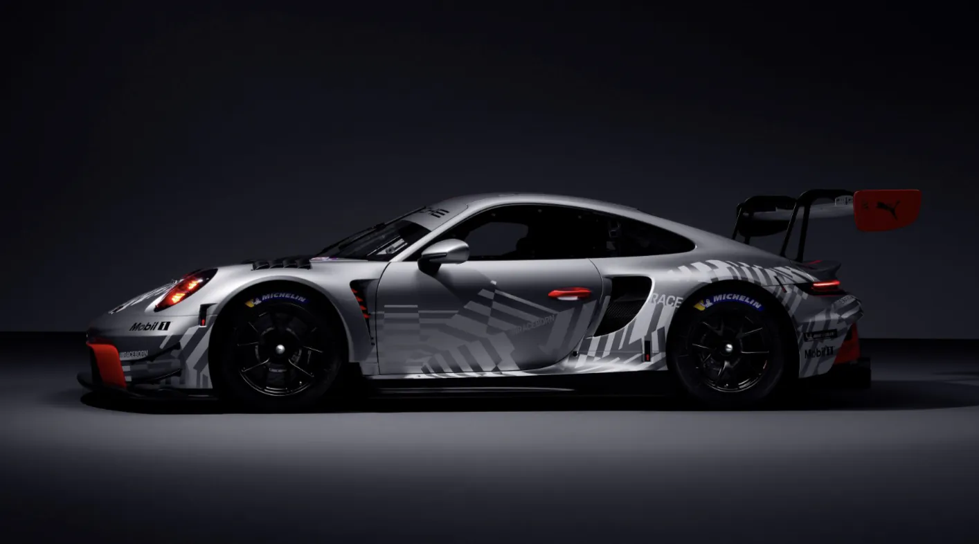 Side profile of a silver Porsche 911 race car with grey geometric decals, red accents, large rear wing, and Michelin tires