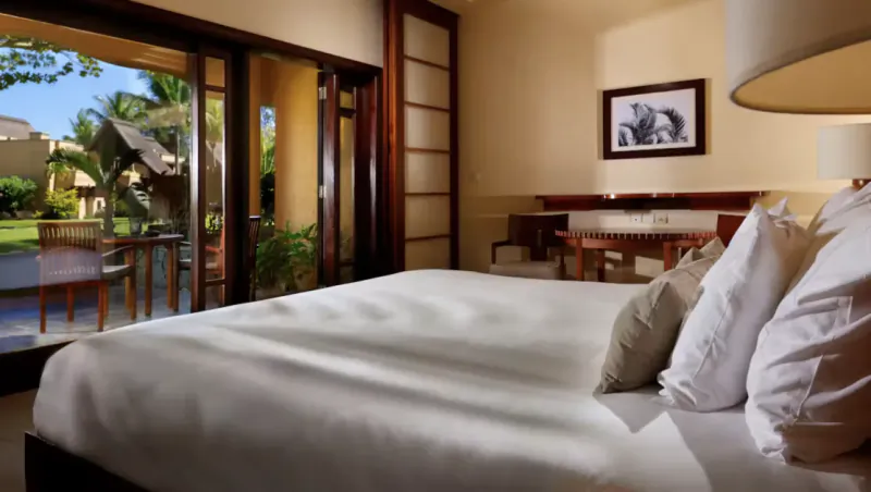 Bright hotel bedroom with large white bed, open balcony doors overlooking tropical garden and patio furniture