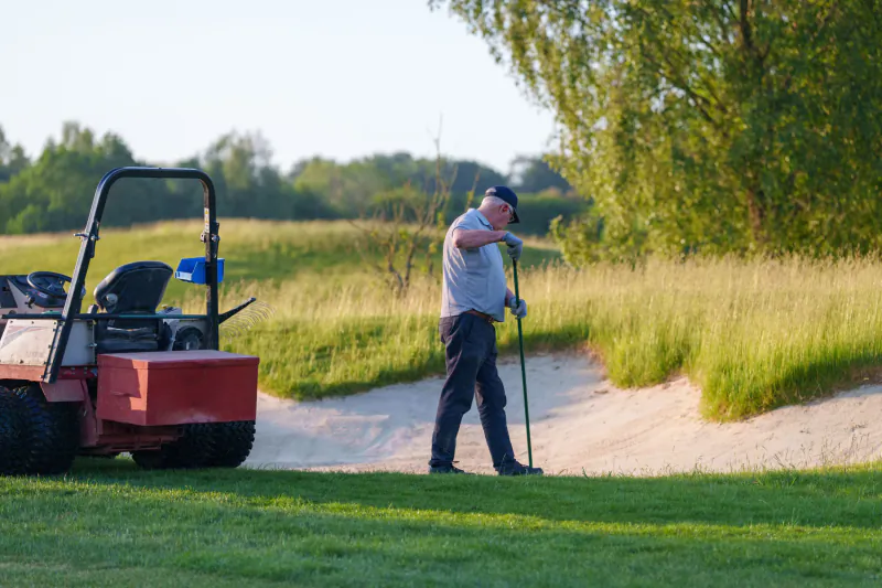 Man in cap and polo shirt holding golf club on sand bunker beside red utility vehicle on golf course