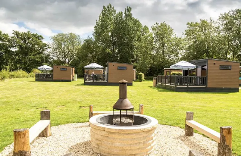 Fire pit with wooden benches overlooking three glamping pods on a grassy field