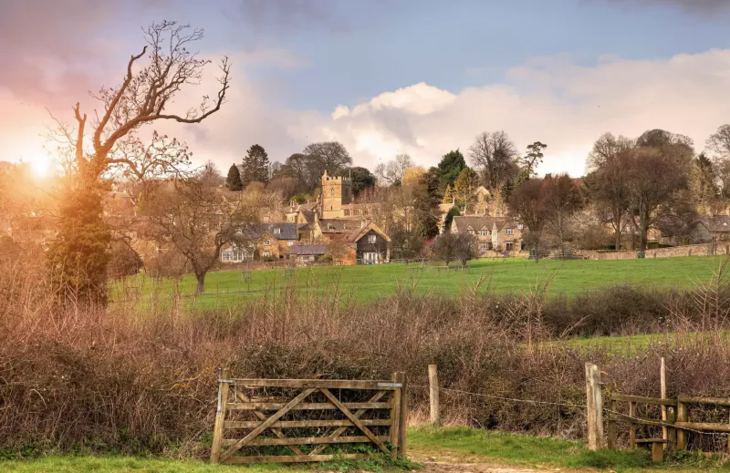 Rural English village with stone buildings and church tower behind a wooden gate and fields at sunset
