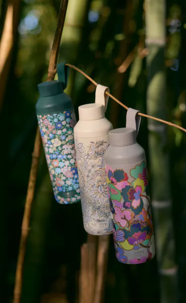 Three colorful floral-patterned water bottles hanging from a rope in a bamboo forest
