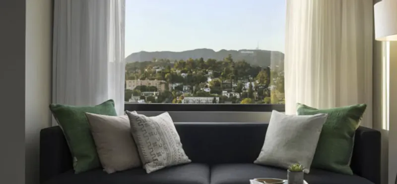 Dark grey sofa with green and beige pillows in front of a large window overlooking Hollywood hills and city skyline