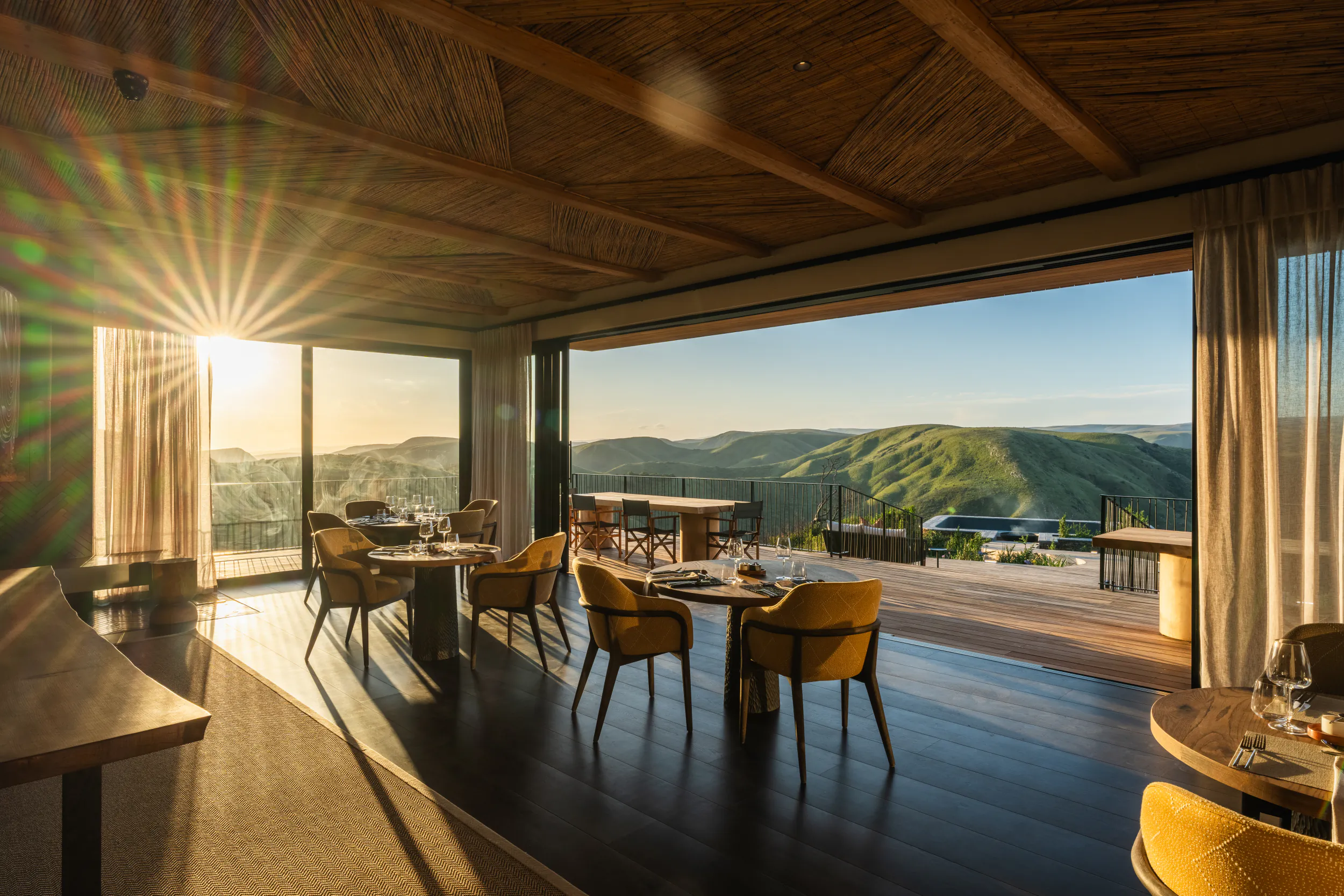 Sunlit luxury restaurant interior with yellow chairs, dark wood floors, and panoramic mountain views at sunset