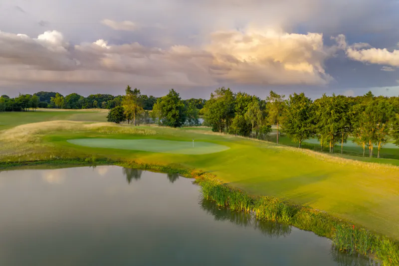 Aerial view of a lush golf course green with a water hazard in the foreground and trees under a dramatic cloudy sky