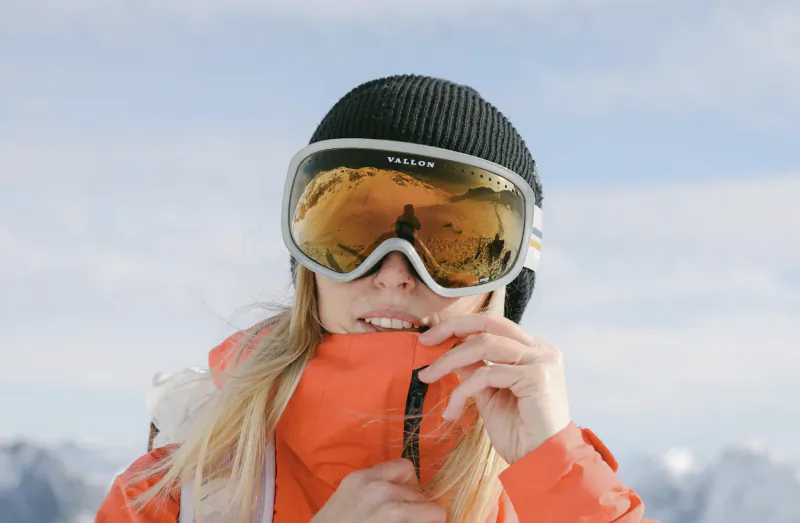 Woman in orange jacket and black beanie adjusting collar, wearing reflective VALLON ski goggles