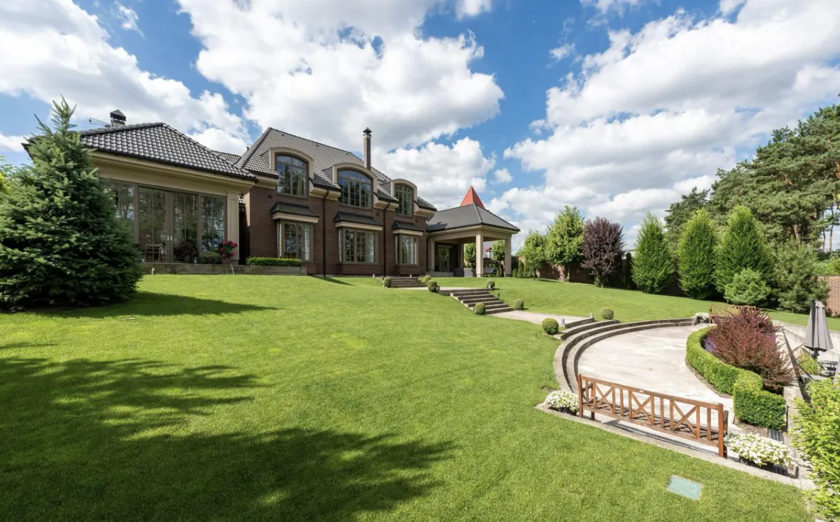 Large two-story brick mansion with arched windows and a red-tiled turret, set on a lush green lawn with a curved stone patio and wooden bench
