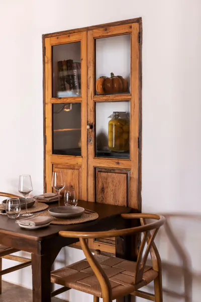Rustic wooden dining table set with plates and wine glasses beside a glass-fronted cabinet displaying books, pumpkins, and a jar of olives