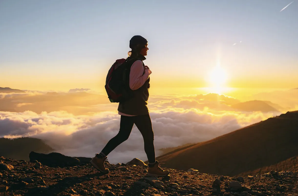 Woman hiking at sunrise with backpack on mountain ridge above clouds