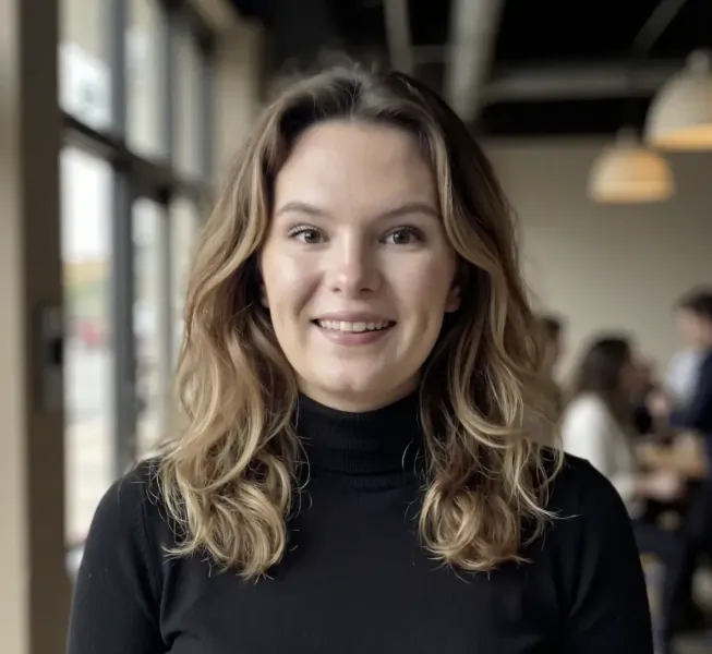 Smiling woman with wavy blonde hair wearing a black turtleneck in a modern office setting