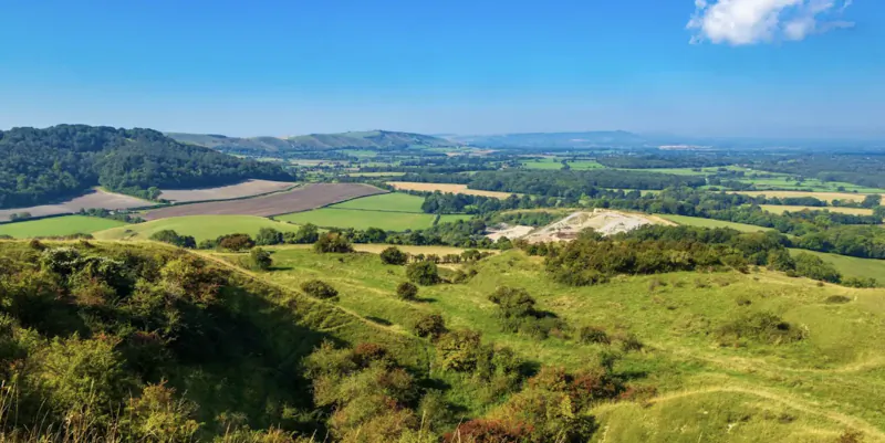 Panoramic view of rolling green hills and patchwork fields under a bright blue sky near Moreton-in-Marsh