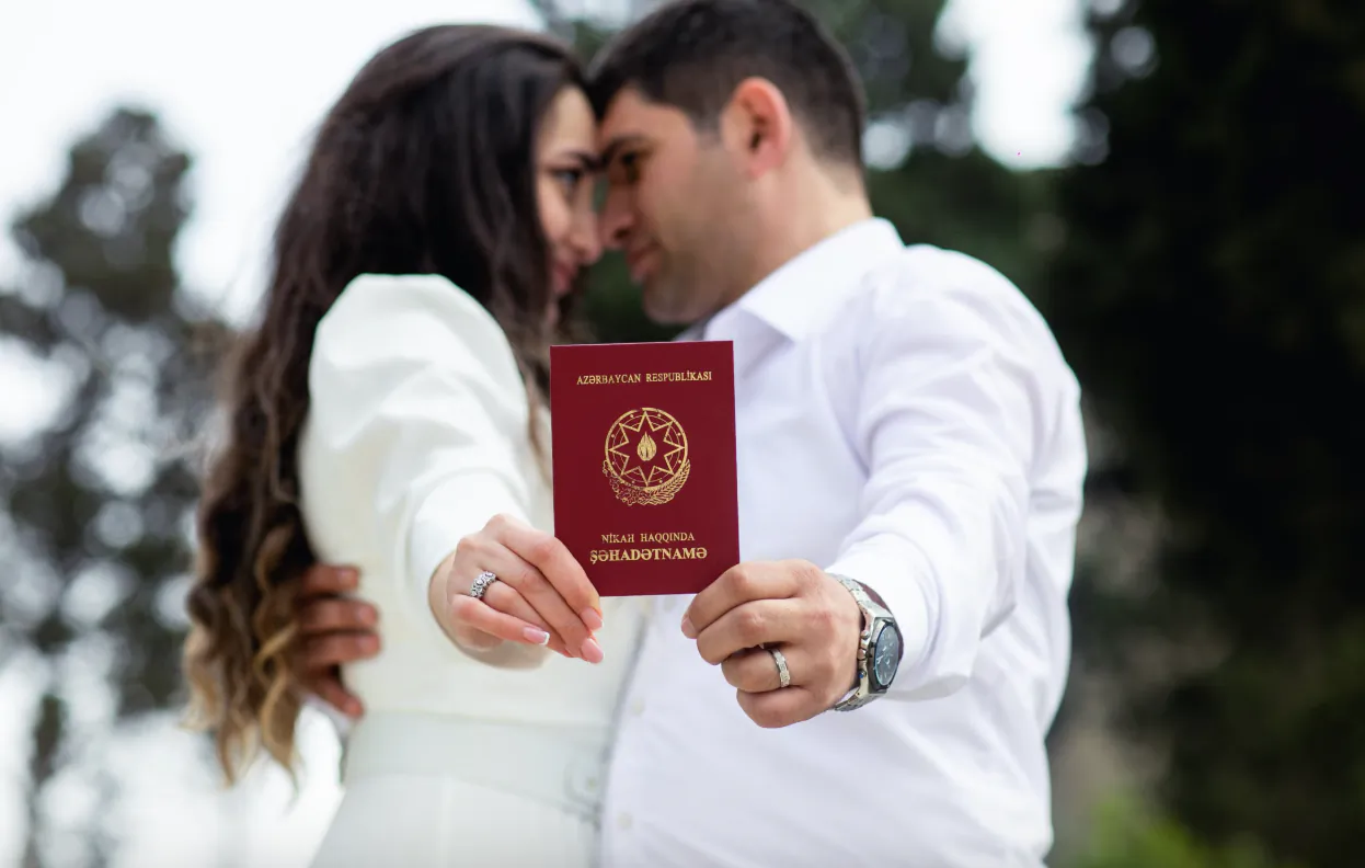 Couple in white attire holding a red Azerbaijani passport together, foreheads touching outdoors
