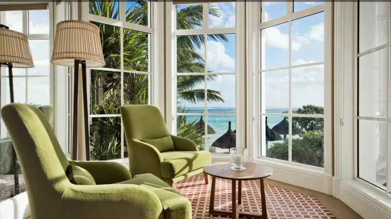 Two green armchairs and a round table in a bright room with large windows overlooking the ocean and palm trees