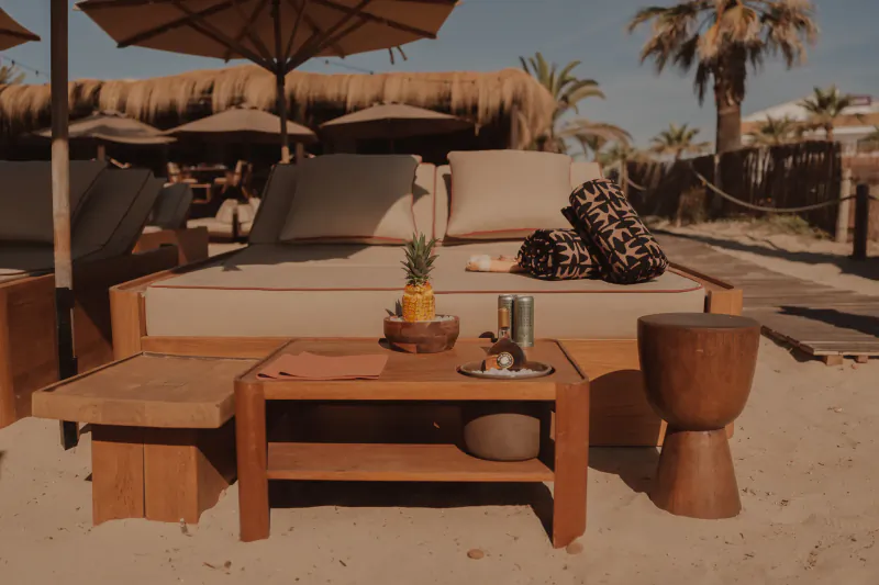Beachside daybed with beige cushions, pineapple and drinks on wooden table, umbrellas and palm trees in background