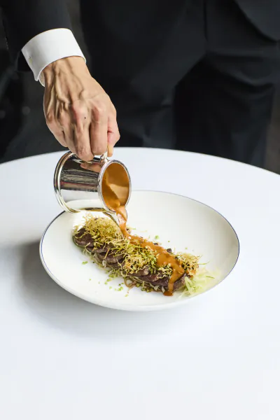 Chef in black suit pours brown sauce from metal pitcher over plated meat dish with microgreens on white table