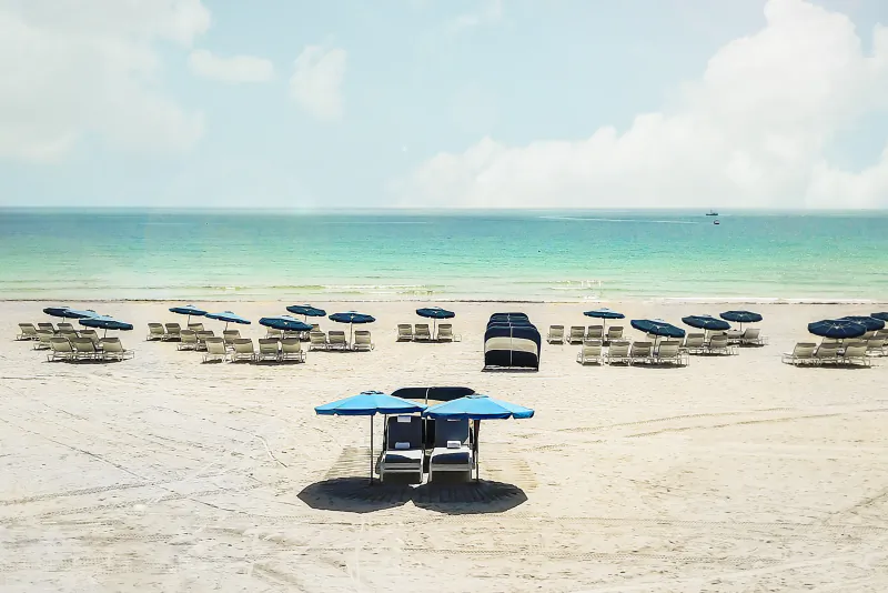 Empty beach with rows of blue umbrellas and lounge chairs facing turquoise ocean under bright sky
