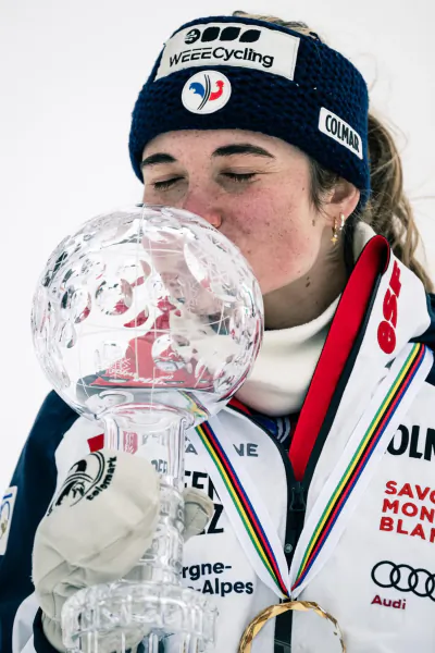 Female skier in French team jacket kissing crystal globe trophy, wearing navy headband with 3000 WEEECycling logo and Colmar patch
