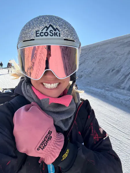 Smiling woman in pink goggles and EcoSki helmet giving a thumbs-up on a snowy slope