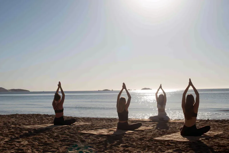 Four women in yoga poses on a sunny beach at sunrise, arms raised overhead facing the sea